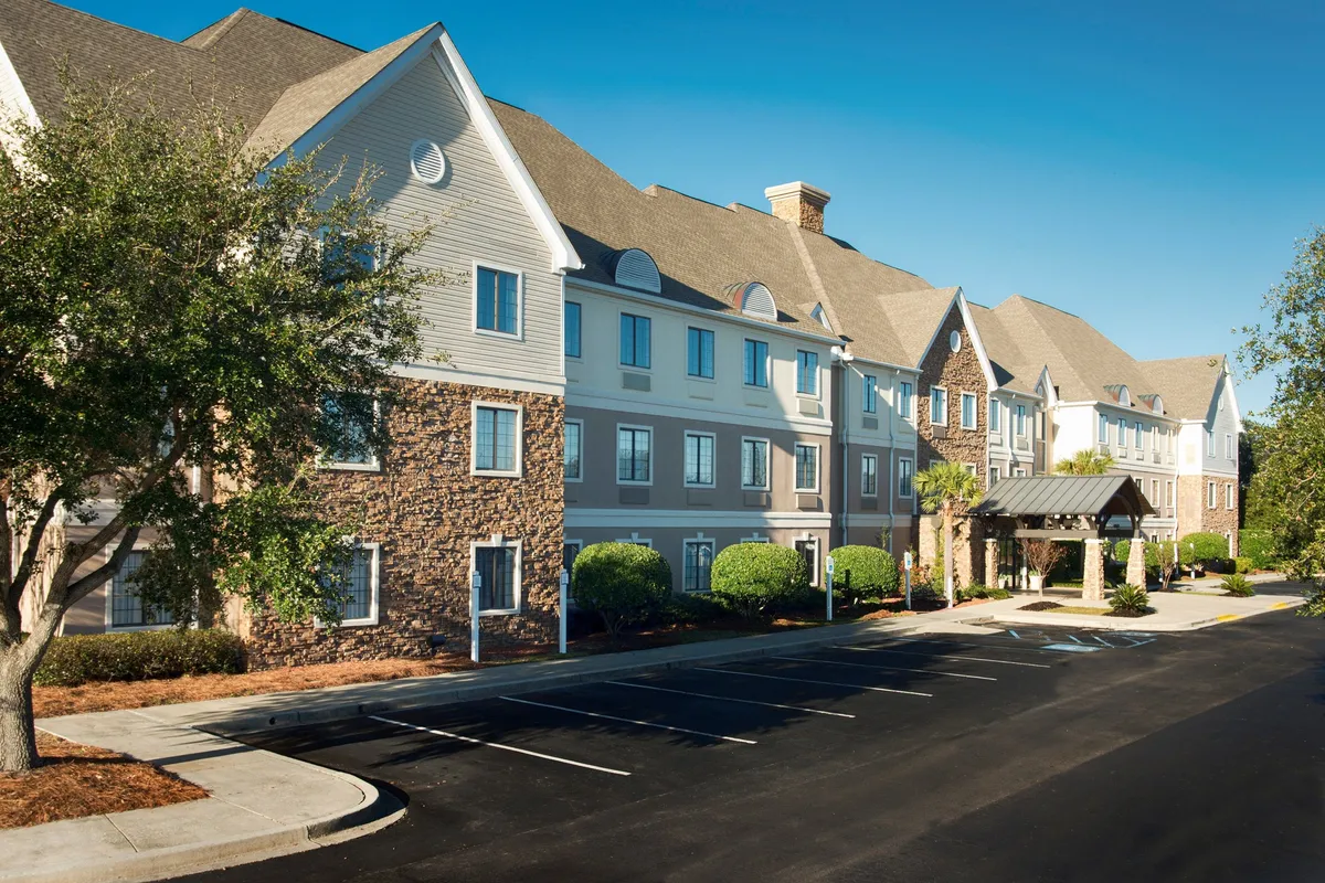 This image captures the stone facade and entrance awning of Staybridge Suites, framed by landscaped greenery. The scene is welcoming and highlights the property's approachable exterior, suggesting a sense of comfort and security for families arriving for their vacation.