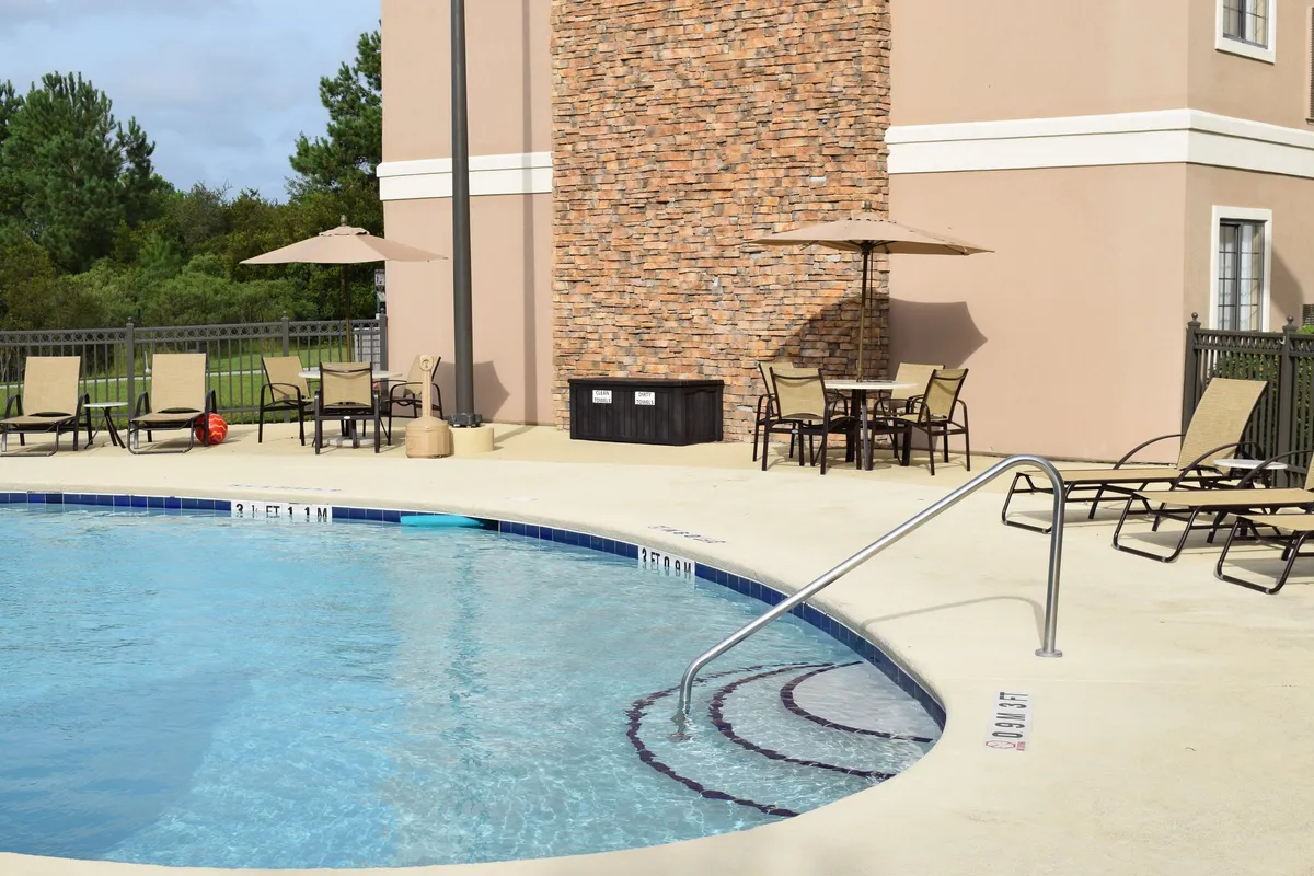 A vibrant outdoor pool scene with sparkling water, surrounded by lounge chairs and umbrellas, set against a warm sunset sky. The area features a stone wall and groups of guests enjoying swimming, sunbathing, and socializing.