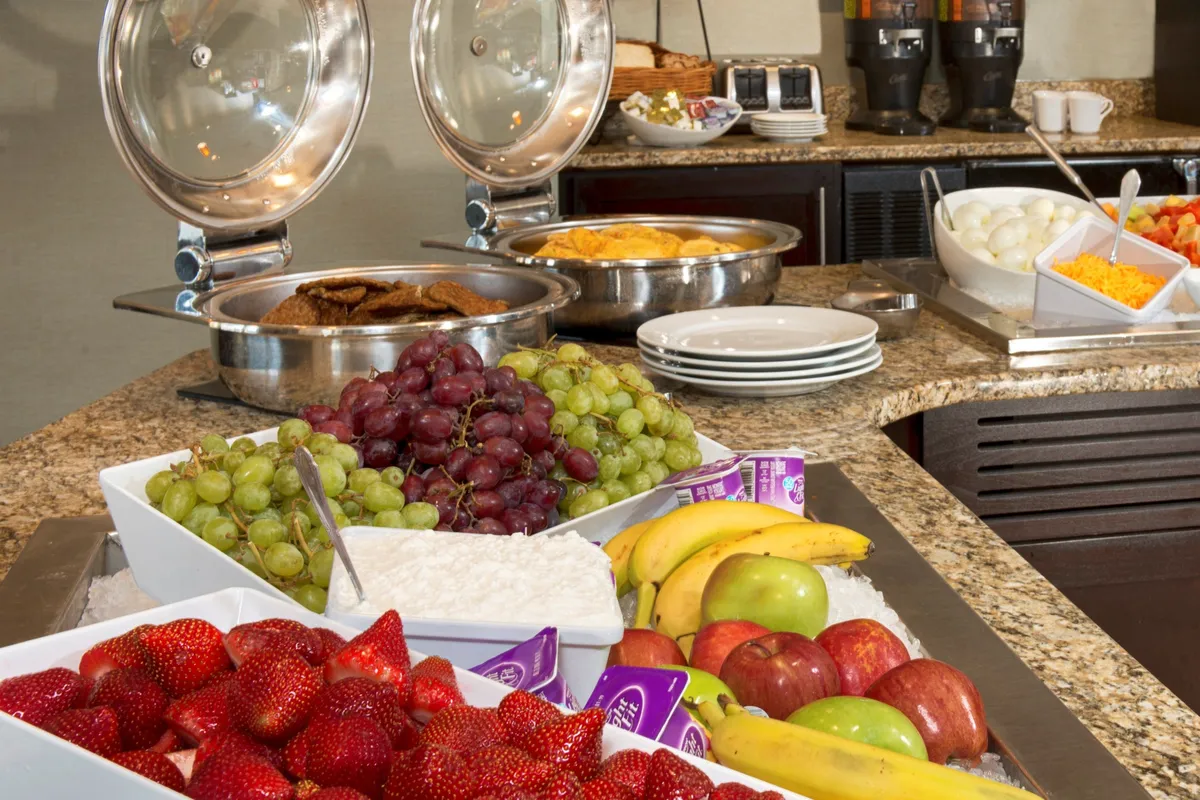 The image displays a selection of fresh fruits, classic breakfast items, coffee machines, and serving dishes, all arranged on a well-organized counter. The mood is relaxed and cheerful, making it easy to imagine guests assembling breakfast or snacks before heading out.