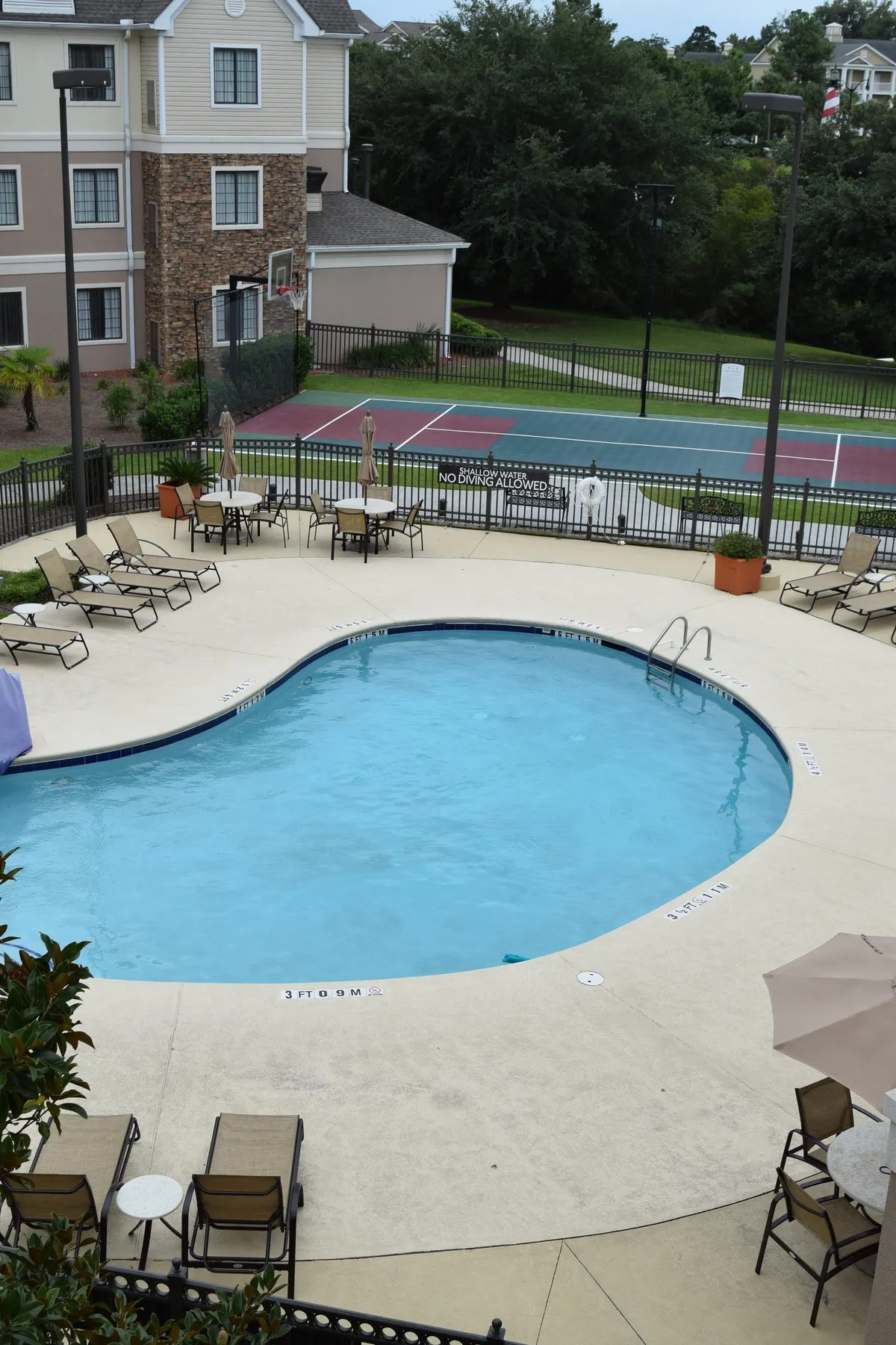 This exterior image captures the swimming pool surrounded by inviting lounge chairs, with a basketball hoop and tennis court visible in the background. The scene is set at sunset, creating a warm, relaxed atmosphere perfect for unwinding after a productive day.