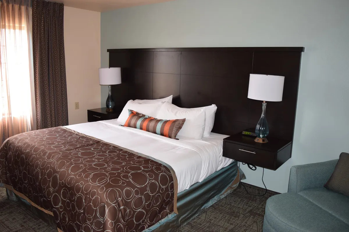 This image features a plush king-sized bed adorned with decorative pillows and flanked by nightstands topped with lamps. An inviting armchair sits by the window, suggesting a cozy reading nook, while soft natural light fills the room, enhancing a peaceful and restful atmosphere.