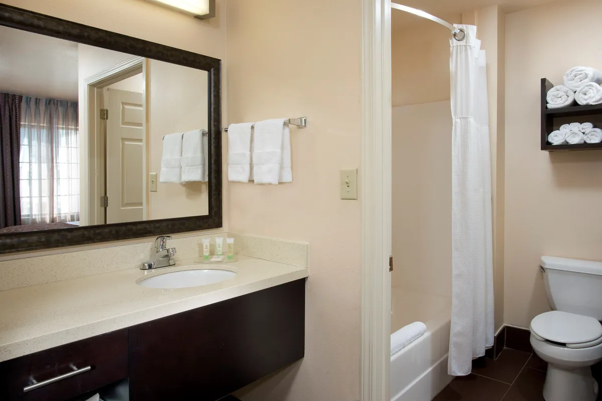 A modern, tidy bathroom is shown with a sleek vanity, a large mirror, and a shower area. Fluffy towels are neatly rolled on a shelf, emphasizing cleanliness and thoughtful amenities for guest comfort.
