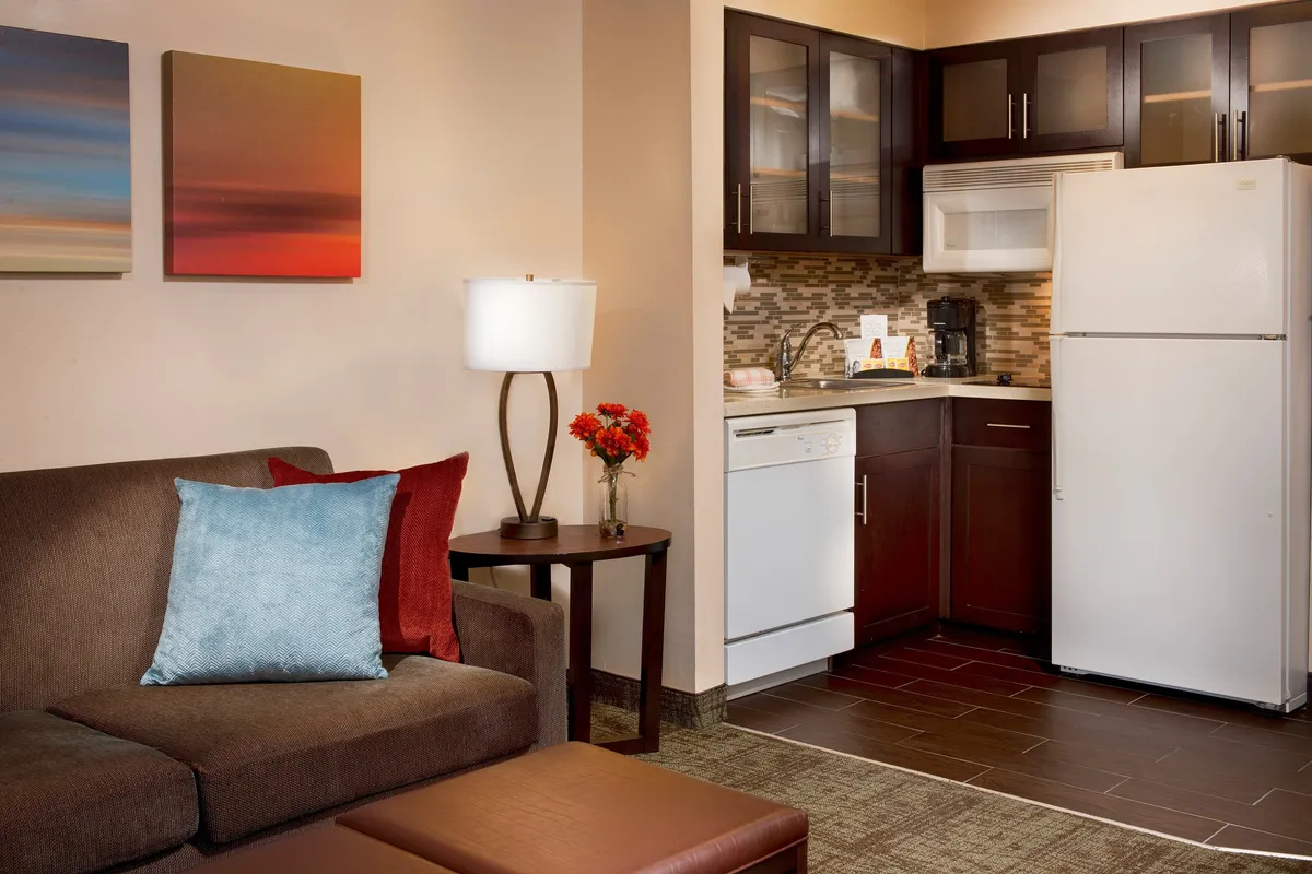 A cozy living room scene featuring a comfortable sofa, a kitchen area in the background, tasteful artwork on the wall, a soft-lit lamp, and a decorative flower arrangement. The layout feels welcoming and is designed for both relaxation and socializing.