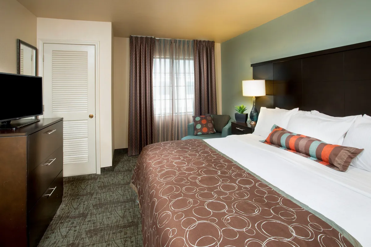 A queen bed dressed with plush, decorative pillows is the focal point of this room. The space includes a dressing table with a mirror and a TV on the dresser, with curtains letting in gentle natural light. The overall vibe is warm and welcoming, ideal for relaxation at any time of day.