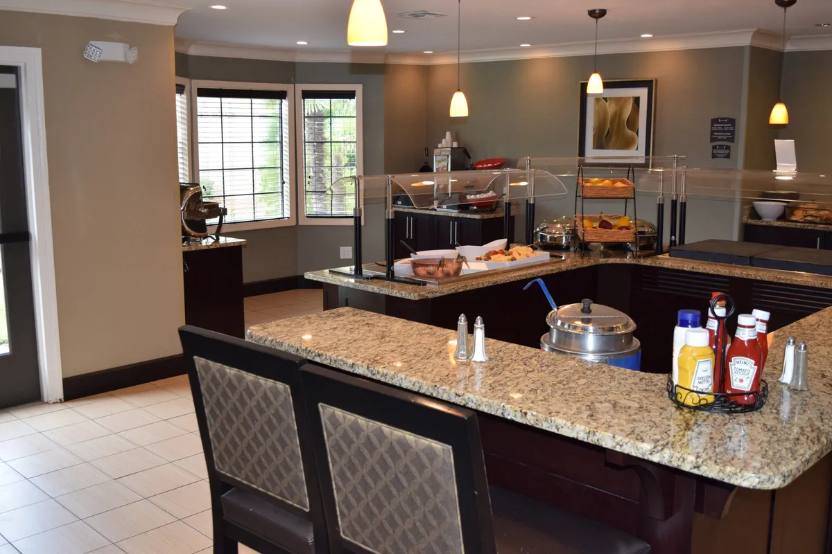 A bright, open kitchen space with a granite countertop, inviting food display, and bar stools. The setup suggests scenes of socializing over breakfast, sharing coffee, or enjoying snacks, with natural light flooding the area.